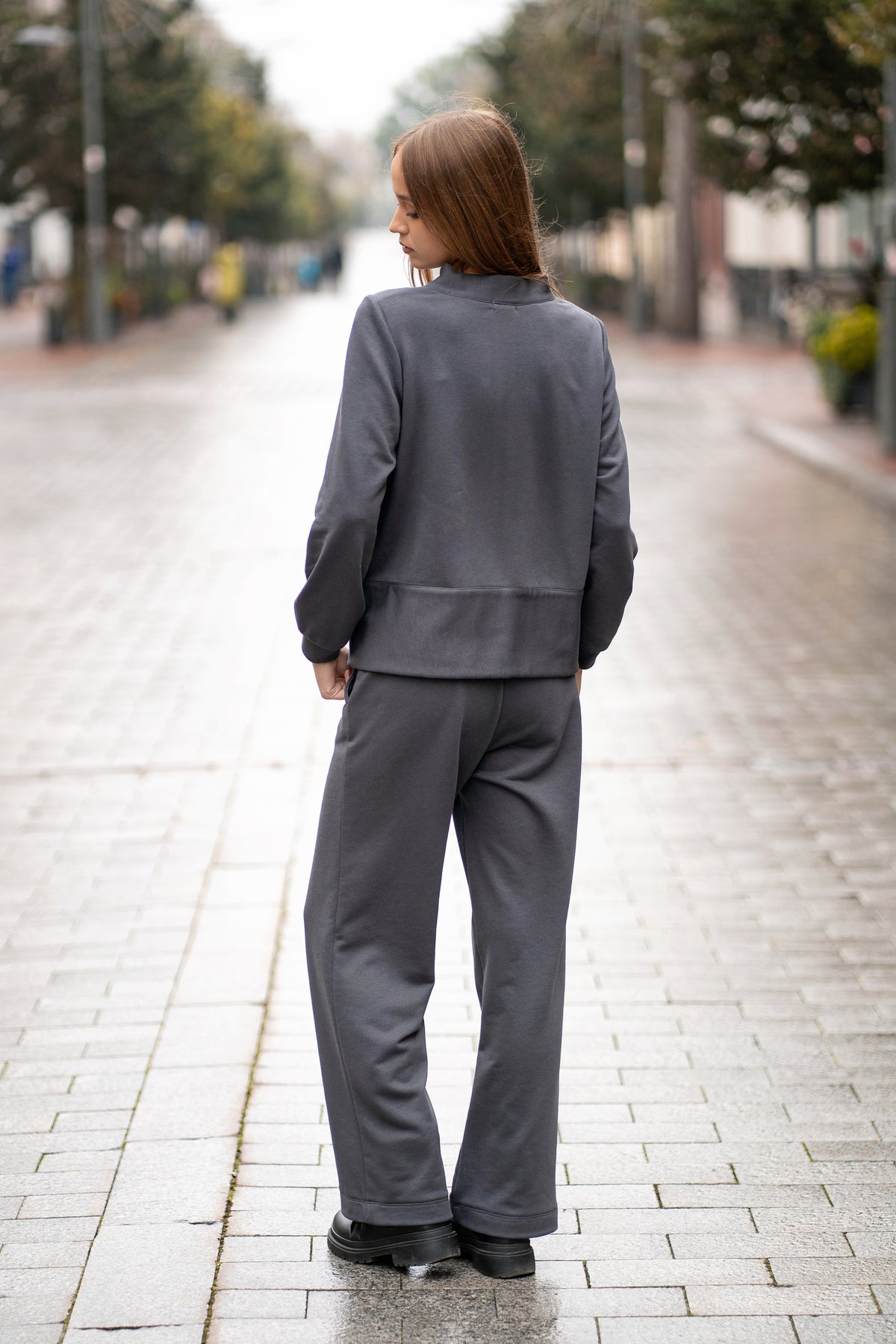Pilkos spalvos trikotažinis kostiumas iš nugaros – džemperis su juodais akcentais ir plačios kelnės, matomas siluetas iš galo.
Back view of the grey women’s jersey set — zip sweatshirt with black trims and wide-leg pants, showing the silhouette from behind.