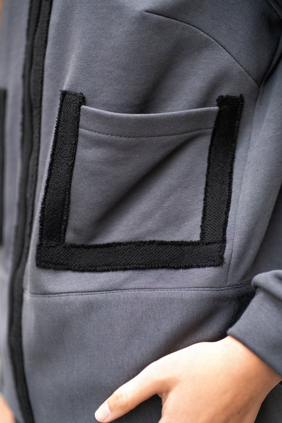 Pilko džemperio detalė iš arti – užtrauktukas ir juodi akcentai prie kišenių, tvarkingas minimalistinis siluetas.
Close-up of the grey zip sweatshirt showing black trims around the zipper and pock