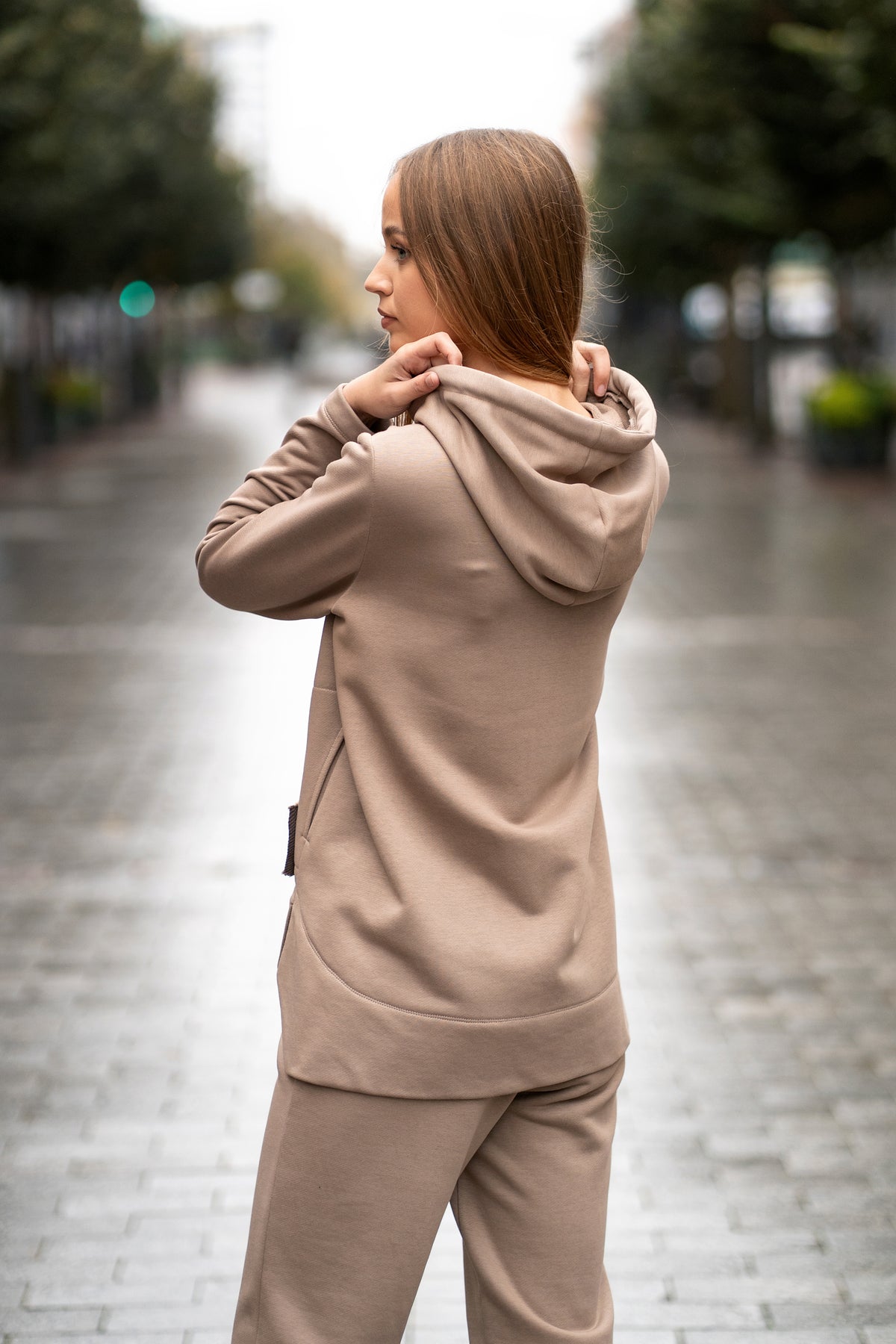 Woman wearing a cappuccino-colored two-piece leisure set with a hood and zipper. The sweatshirt has an asymmetric cut, front pockets, and a hand-decorated face motif. The trousers are straight and comfortable with an elastic waistband and pockets. Modern, cozy, and stylish outfit for everyday wear. / Moteriškas kapučino spalvos kostiumas su gobtuvu, dekoru ir kišenėmis – stilingas ir patogus kasdienai.