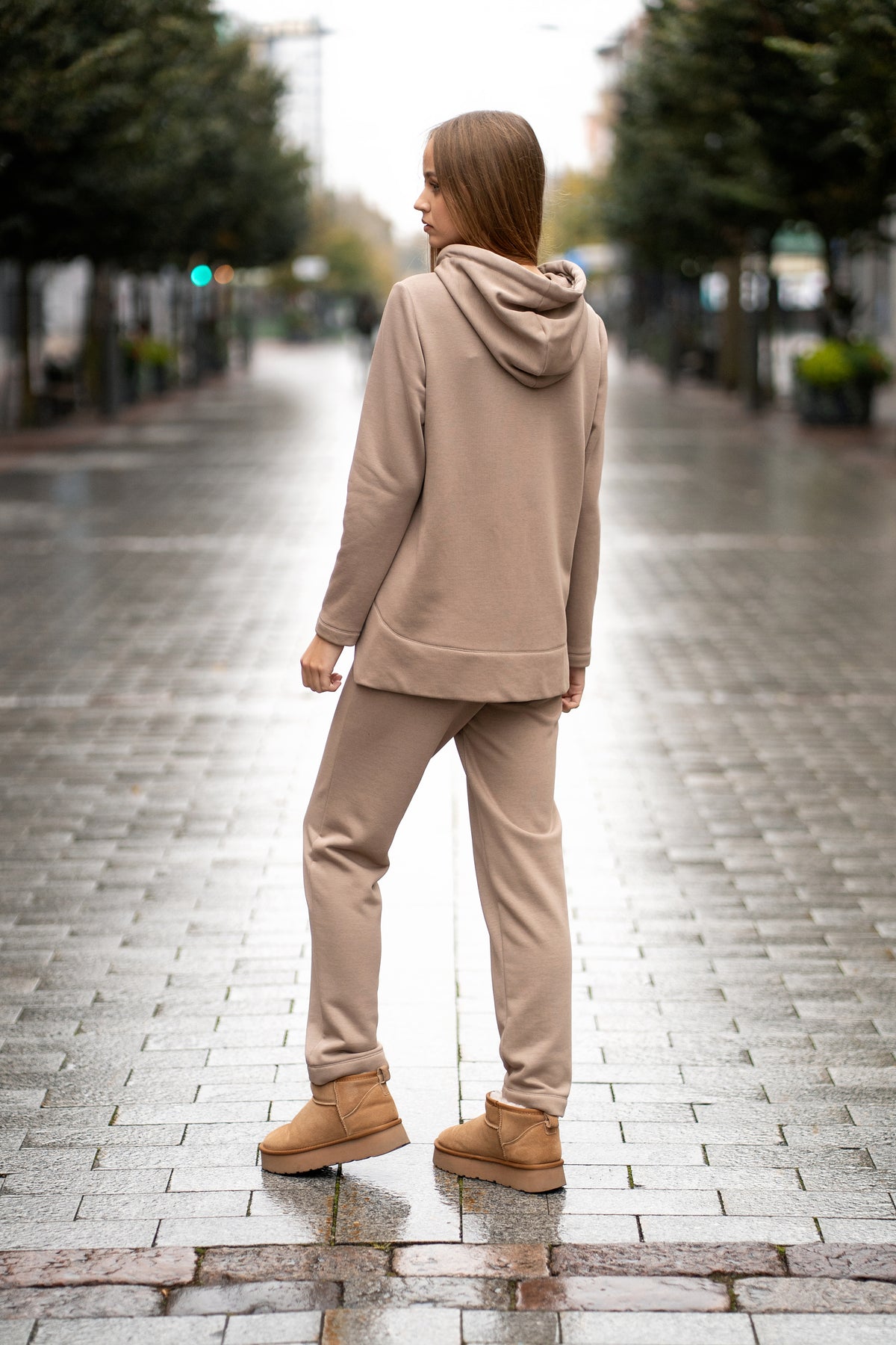 Woman wearing a cappuccino-colored two-piece leisure set with a hood and zipper. The sweatshirt has an asymmetric cut, front pockets, and a hand-decorated face motif. The trousers are straight and comfortable with an elastic waistband and pockets. Modern, cozy, and stylish outfit for everyday wear. / Moteriškas kapučino spalvos kostiumas su gobtuvu, dekoru ir kišenėmis – stilingas ir patogus kasdienai.