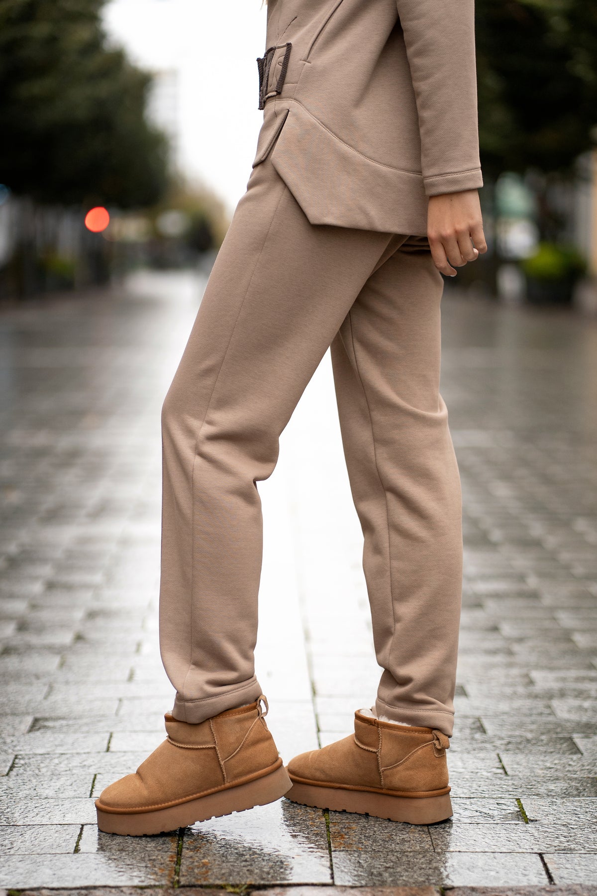 Woman wearing a cappuccino-colored two-piece leisure set with a hood and zipper. The sweatshirt has an asymmetric cut, front pockets, and a hand-decorated face motif. The trousers are straight and comfortable with an elastic waistband and pockets. Modern, cozy, and stylish outfit for everyday wear. / Moteriškas kapučino spalvos kostiumas su gobtuvu, dekoru ir kišenėmis – stilingas ir patogus kasdienai.