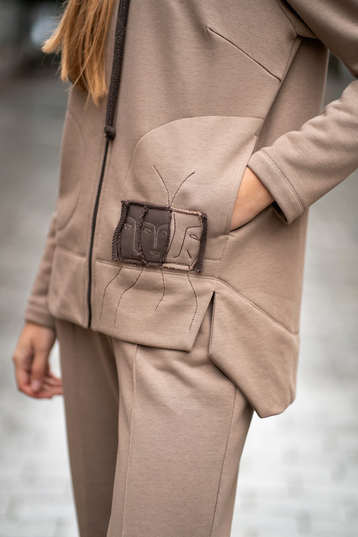 Woman wearing a cappuccino-colored two-piece leisure set with a hood and zipper. The sweatshirt has an asymmetric cut, front pockets, and a hand-decorated face motif. The trousers are straight and comfortable with an elastic waistband and pockets. Modern, cozy, and stylish outfit for everyday wear. / Moteriškas kapučino spalvos kostiumas su gobtuvu, dekoru ir kišenėmis – stilingas ir patogus kasdienai.