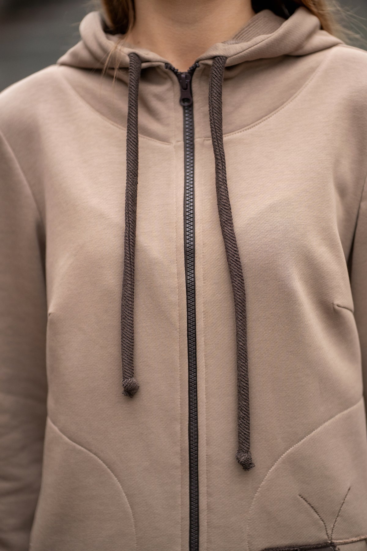 Woman wearing a cappuccino-colored two-piece leisure set with a hood and zipper. The sweatshirt has an asymmetric cut, front pockets, and a hand-decorated face motif. The trousers are straight and comfortable with an elastic waistband and pockets. Modern, cozy, and stylish outfit for everyday wear. / Moteriškas kapučino spalvos kostiumas su gobtuvu, dekoru ir kišenėmis – stilingas ir patogus kasdienai.