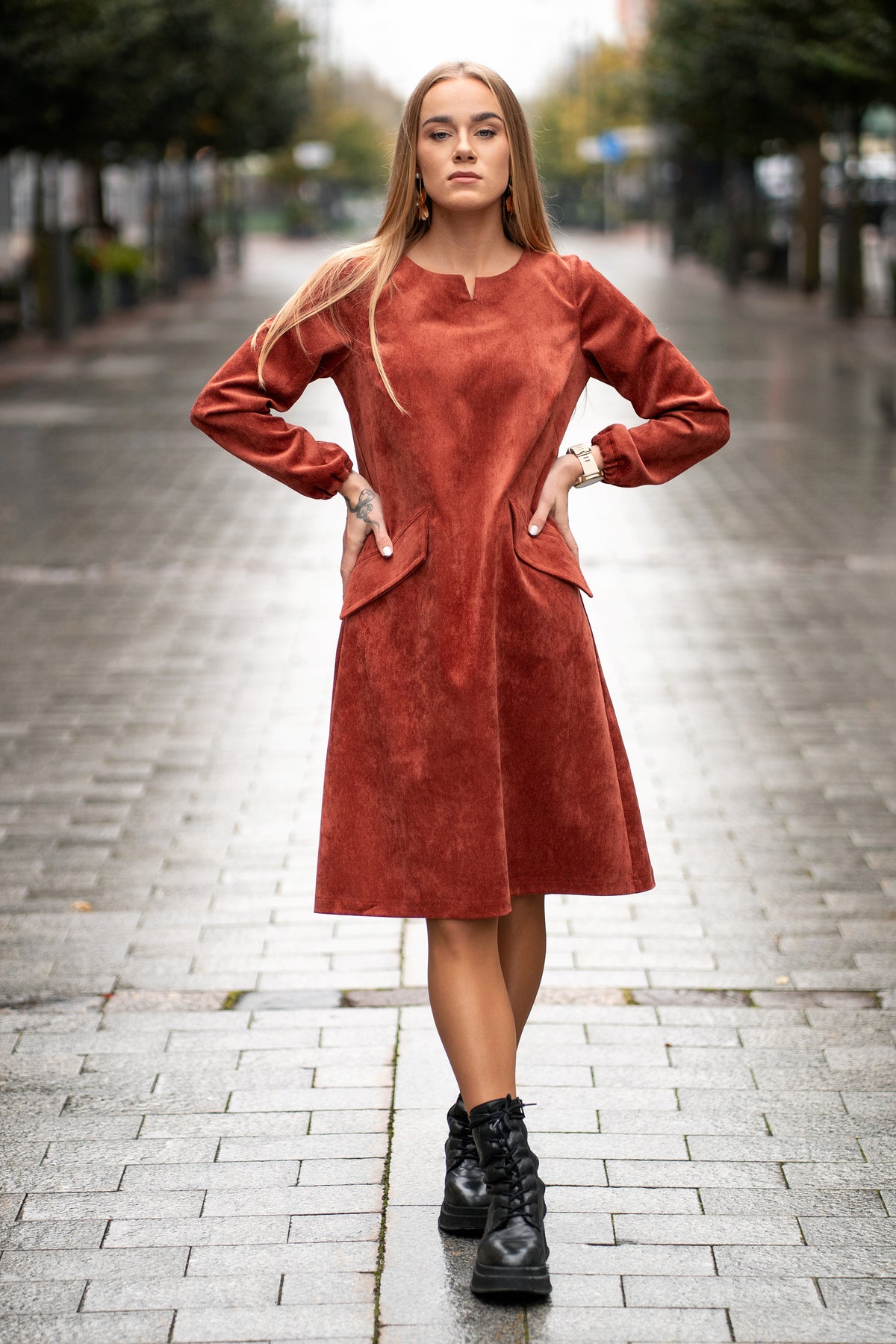 Woman wearing a terracotta velvet (corduroy) dress with long sleeves gathered at the cuffs, side pockets, and a subtle neckline detail. The dress is made from a soft cotton-based fabric with fine stripes. Mid-length, breathable and elegant design for autumn and winter. / Terakotos spalvos veliūrinė suknelė su kišenėmis ir subtiliu kaklo dizainu, iš medvilninio audinio su juostelėmis.