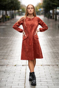 Woman wearing a terracotta velvet (corduroy) dress with long sleeves gathered at the cuffs, side pockets, and a subtle neckline detail. The dress is made from a soft cotton-based fabric with fine stripes. Mid-length, breathable and elegant design for autumn and winter. / Terakotos spalvos veliūrinė suknelė su kišenėmis ir subtiliu kaklo dizainu, iš medvilninio audinio su juostelėmis.