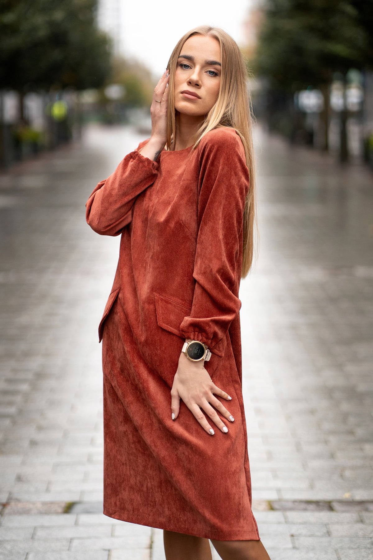 Woman wearing a terracotta velvet (corduroy) dress with long sleeves gathered at the cuffs, side pockets, and a subtle neckline detail. The dress is made from a soft cotton-based fabric with fine stripes. Mid-length, breathable and elegant design for autumn and winter. / Terakotos spalvos veliūrinė suknelė su kišenėmis ir subtiliu kaklo dizainu, iš medvilninio audinio su juostelėmis.