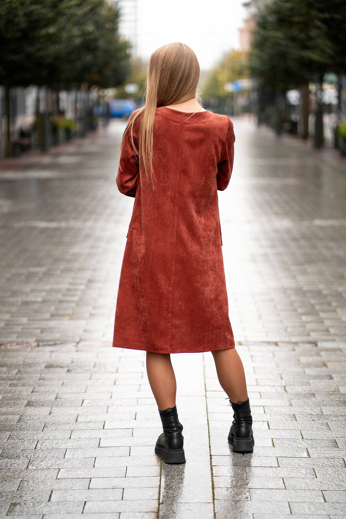 Woman wearing a terracotta velvet (corduroy) dress with long sleeves gathered at the cuffs, side pockets, and a subtle neckline detail. The dress is made from a soft cotton-based fabric with fine stripes. Mid-length, breathable and elegant design for autumn and winter. / Terakotos spalvos veliūrinė suknelė su kišenėmis ir subtiliu kaklo dizainu, iš medvilninio audinio su juostelėmis.