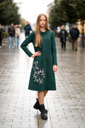 Woman wearing a dark green cotton jersey dress with a bell silhouette. The dress features long sleeves, side pockets, and handmade floral embroidery on the side. Elegant and comfortable mid-weight fabric, ideal for everyday wear or special occasions. / Žalios spalvos trikotažinė varpelio silueto suknelė su šoniniu rankų darbo gėlių dekoru.