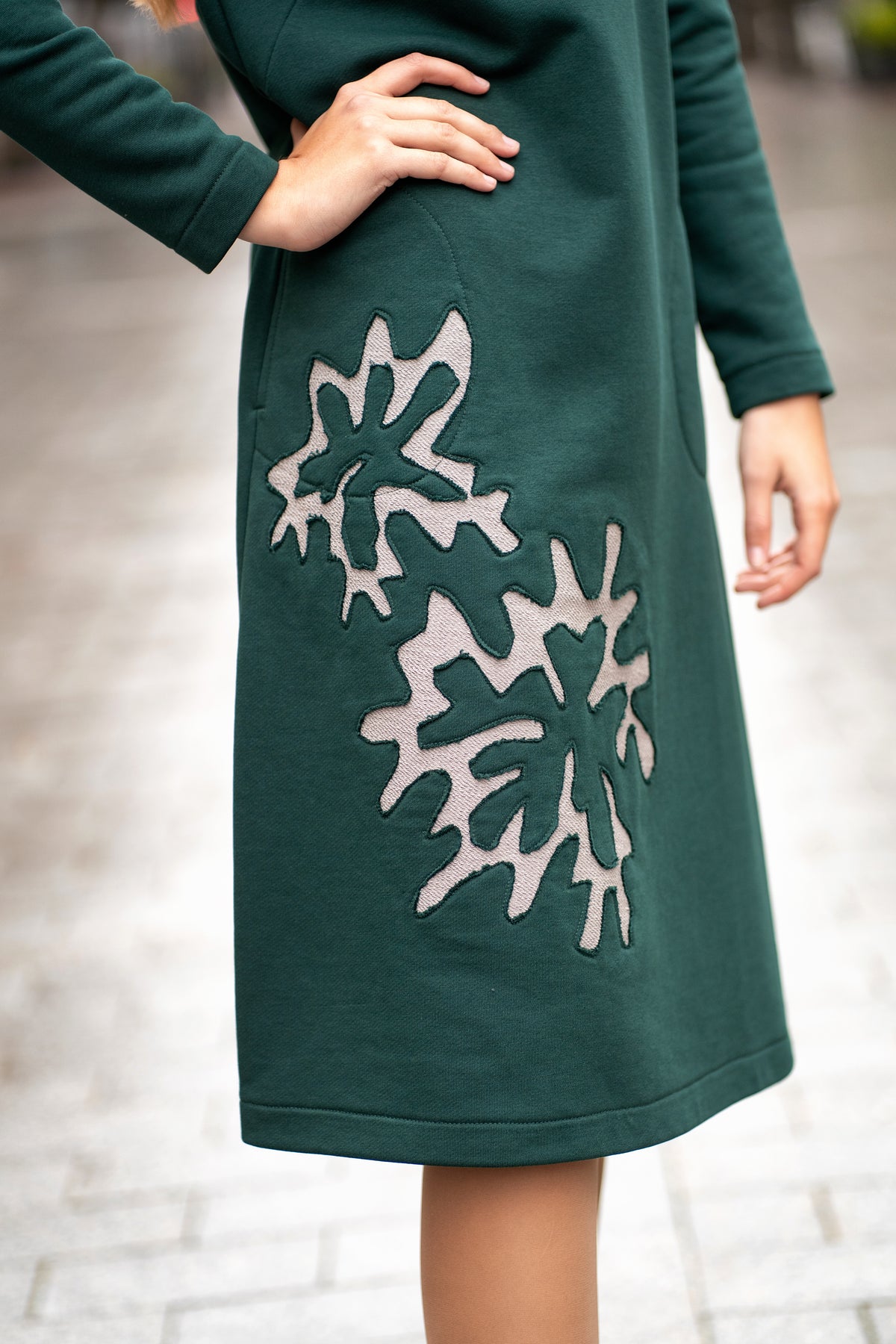 Woman wearing a dark green cotton jersey dress with a bell silhouette. The dress features long sleeves, side pockets, and handmade floral embroidery on the side. Elegant and comfortable mid-weight fabric, ideal for everyday wear or special occasions. / Žalios spalvos trikotažinė varpelio silueto suknelė su šoniniu rankų darbo gėlių dekoru.