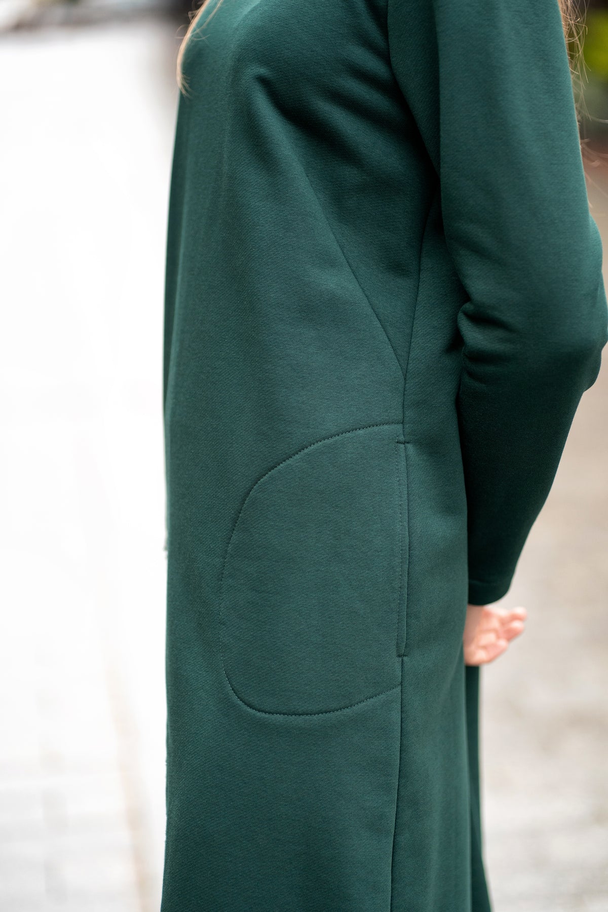 Woman wearing a dark green cotton jersey dress with a bell silhouette. The dress features long sleeves, side pockets, and handmade floral embroidery on the side. Elegant and comfortable mid-weight fabric, ideal for everyday wear or special occasions. / Žalios spalvos trikotažinė varpelio silueto suknelė su šoniniu rankų darbo gėlių dekoru.