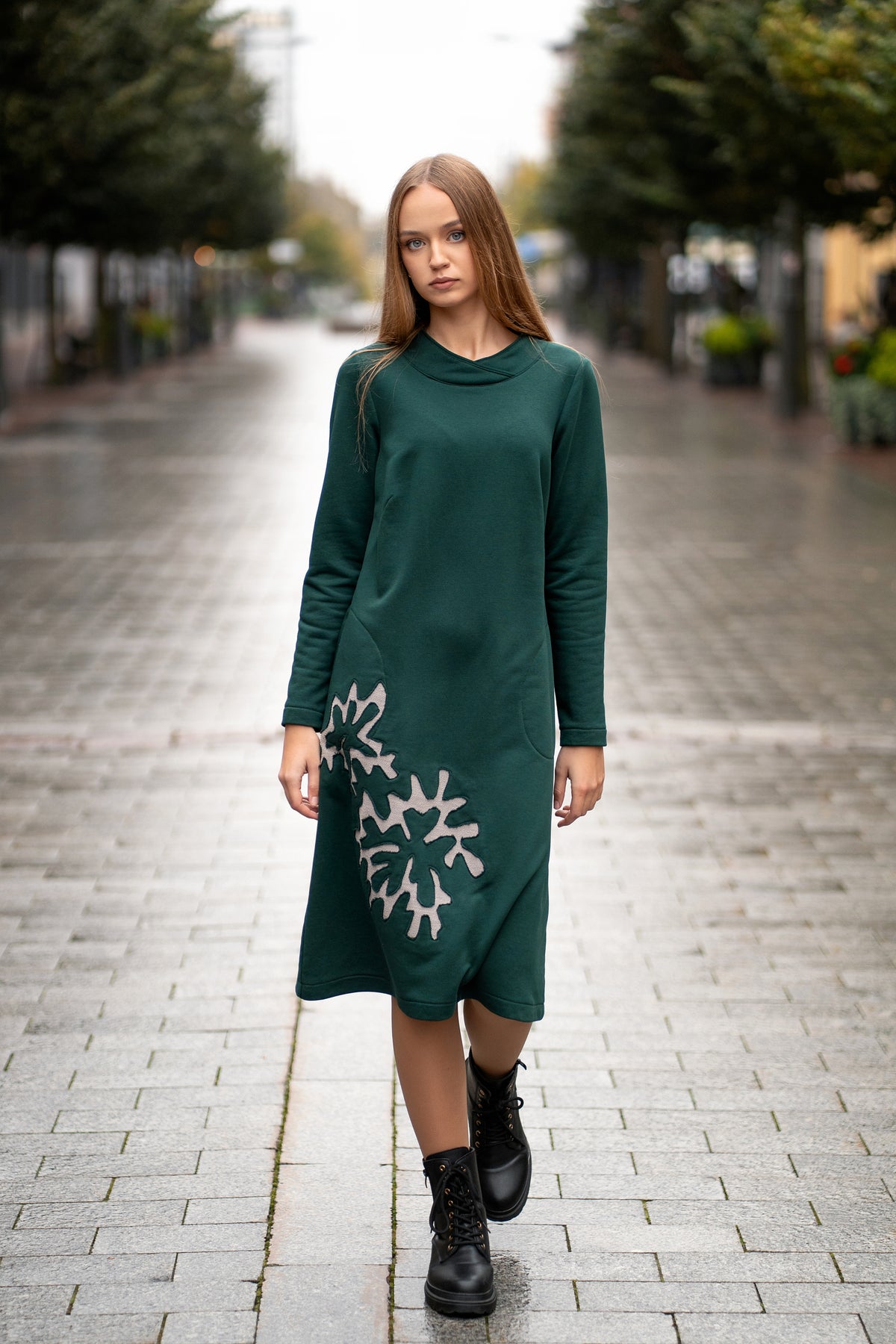 Woman wearing a dark green cotton jersey dress with a bell silhouette. The dress features long sleeves, side pockets, and handmade floral embroidery on the side. Elegant and comfortable mid-weight fabric, ideal for everyday wear or special occasions. / Žalios spalvos trikotažinė varpelio silueto suknelė su šoniniu rankų darbo gėlių dekoru.