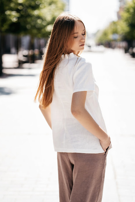 Balta lininė palaidinė moterims – trumpomis rankovėmis, dekoruota subtiliomis klostelėmis aplink kakliuką ir per priekį, siūta iš minkštinto natūralaus lino – White linen blouse for women with delicate pleated detail and short sleeves, perfect for summer wear.