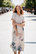 Model wearing a long linen dress with artistic watercolor floral print, made from 100% natural softened linen. The dress features a slit neckline, short sleeves, side pockets, and elegant side slits for comfort. Handmade in Lithuania. Moteriška lininė suknelė su gėlių raštu, siūta iš minkštinto lino, puikiai tinka vasarai.

