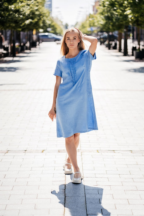 Light blue linen summer dress for women made from 100% softened linen, featuring a relaxed silhouette, short sleeves, two side pockets, and a delicate handmade-like decorative front detail. Ideal for warm weather, breathable and stylish. Modelis vilki melsvą lininę suknelę trumpomis rankovėmis, tinkamą vasarai – natūralus audinys, patogumas ir elegancija viename.
