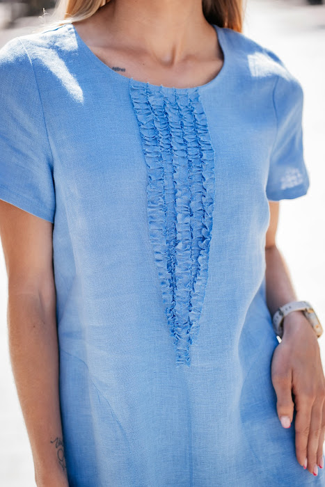 Light blue linen summer dress for women made from 100% softened linen, featuring a relaxed silhouette, short sleeves, two side pockets, and a delicate handmade-like decorative front detail. Ideal for warm weather, breathable and stylish. Modelis vilki melsvą lininę suknelę trumpomis rankovėmis, tinkamą vasarai – natūralus audinys, patogumas ir elegancija viename.