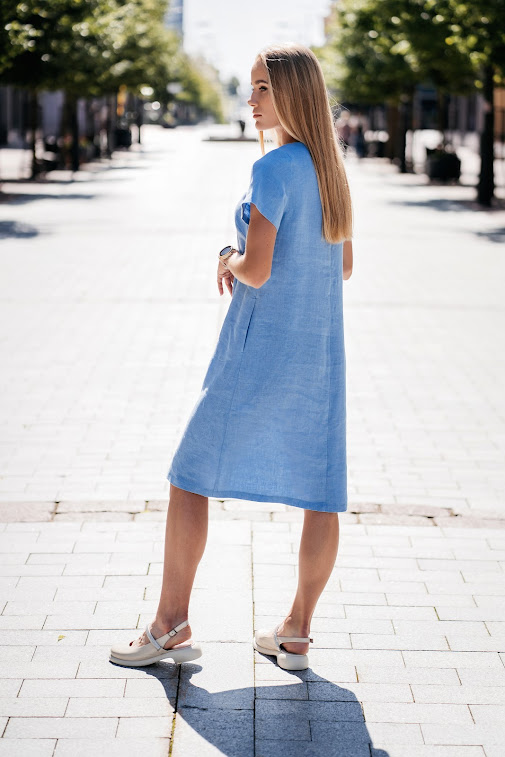 Light blue linen summer dress for women made from 100% softened linen, featuring a relaxed silhouette, short sleeves, two side pockets, and a delicate handmade-like decorative front detail. Ideal for warm weather, breathable and stylish. Modelis vilki melsvą lininę suknelę trumpomis rankovėmis, tinkamą vasarai – natūralus audinys, patogumas ir elegancija viename.
