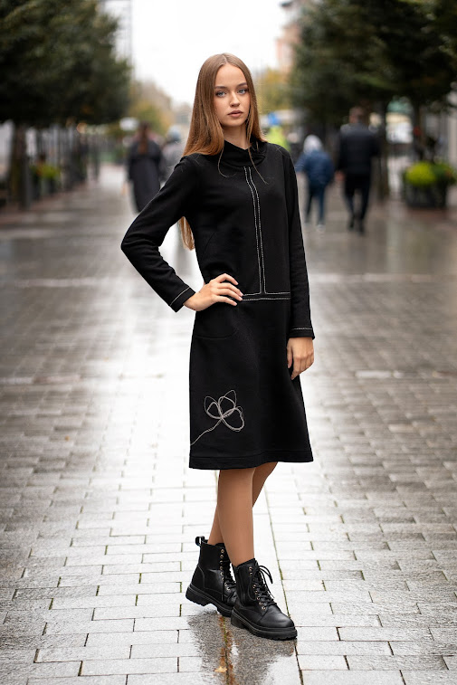Juoda trikotažinė suknelė tiesaus silueto, su dekoratyvinėmis siūlėmis ir rankų darbo aplikacija, modelis stovi miesto gatvėje – black knit dress with decorative seams, handmade applique, straight silhouett