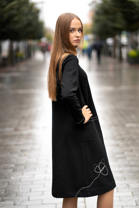 Juoda trikotažinė suknelė tiesaus silueto, su dekoratyvinėmis siūlėmis ir rankų darbo aplikacija, modelis stovi miesto gatvėje – black knit dress with decorative seams, handmade applique, straight silhouett