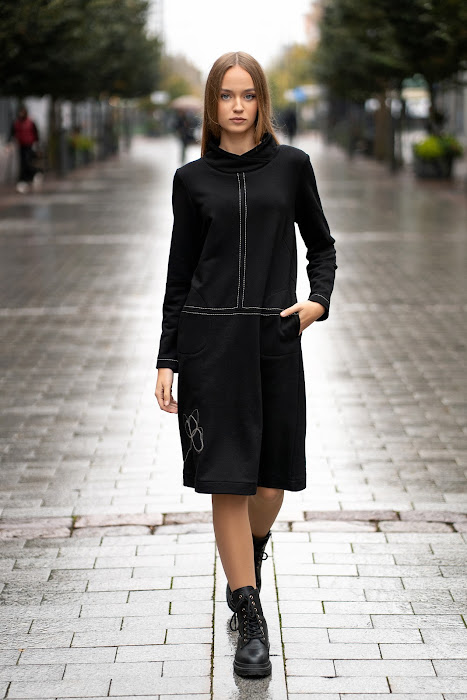 Juoda trikotažinė suknelė tiesaus silueto, su dekoratyvinėmis siūlėmis ir rankų darbo aplikacija, modelis stovi miesto gatvėje – black knit dress with decorative seams, handmade applique, straight silhouett