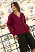 A woman wearing a burgundy V-neck cardigan with buttons and front pockets, made of soft viscose-blend knit, styled with black trousers on a balcony with elegant architectural background. The knit is smooth, cozy, and structured. | Moteris dėvi bordo spalvos kardiganą su sagomis, V formos iškirpte ir kišenėmis, švelnaus ir forma išlaikančio mezgimo, fotografuota ant balkono su miesto architektūra.