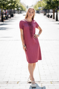 Woman wearing a dark rose linen summer dress with handmade front detail, standing on a sunlit street. Suknelė iš 100 % minkštinto lino, dekoruota priekyje, kišenės šonuose, lengvo silueto, idealiai tinka vasarai. Modelis vilki dydį 36.