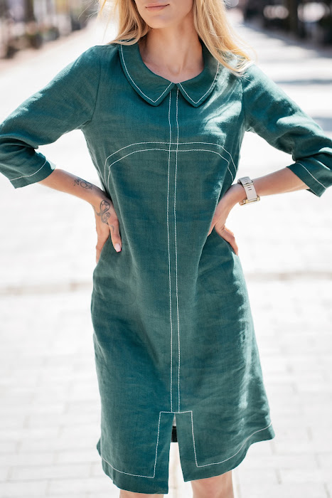 Woman wearing a dark green linen dress with 3/4 sleeves and school-style collar, standing on a sunny street. / Moteris dėvi tamsiai žalią lininę suknelę su 3/4 rankovėmis ir mokykline apykakle, stovi miesto gatvėje.

Close-up of front design showing decorative seams and soft linen fabric. / Artimas vaizdas į priekį – matomos išryškintos siūlės ir minkštas lino audinys.

Side view of the linen dress with subtle silhouette and front slit. / Šoninis vaizdas – subtilus siluetas ir skeltukas priekyje.