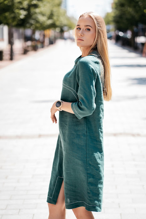 Woman wearing a dark green linen dress with 3/4 sleeves and school-style collar, standing on a sunny street. / Moteris dėvi tamsiai žalią lininę suknelę su 3/4 rankovėmis ir mokykline apykakle, stovi miesto gatvėje.

Close-up of front design showing decorative seams and soft linen fabric. / Artimas vaizdas į priekį – matomos išryškintos siūlės ir minkštas lino audinys.

Side view of the linen dress with subtle silhouette and front slit. / Šoninis vaizdas – subtilus siluetas ir skeltukas priekyje.